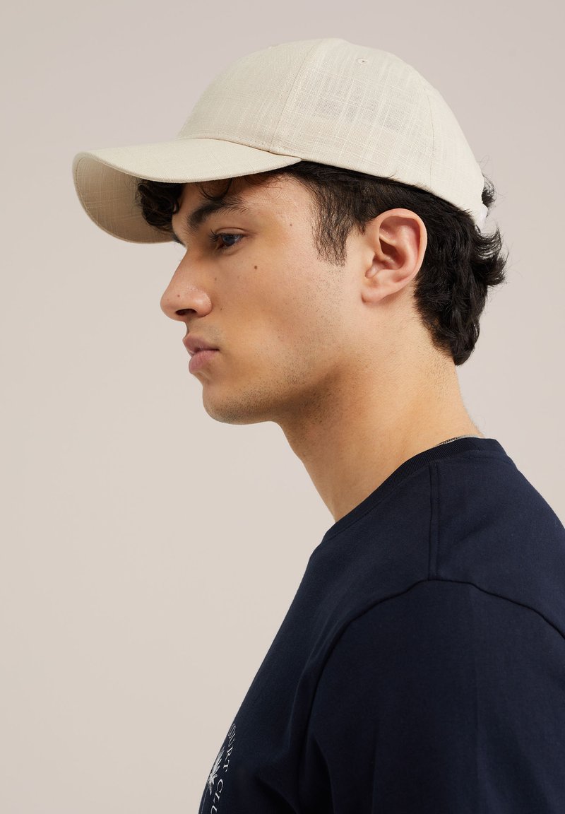Jeune homme portant une casquette beige et une chemise foncée, regardant vers la gauche sur un fond clair uni, vu de profil.