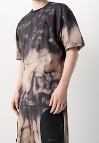 Camiseta de manga corta en negro y tan con efecto tie-dye, que presenta un bolsillo con cremallera en el pecho, cuello redondo y costuras contrastantes.