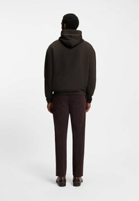 Felpa con cappuccio di colore marrone scuro con stringhe, orlo a costine e vestibilità rilassata, abbinata a pantaloni scuri e scarpe marroni, vista da dietro.