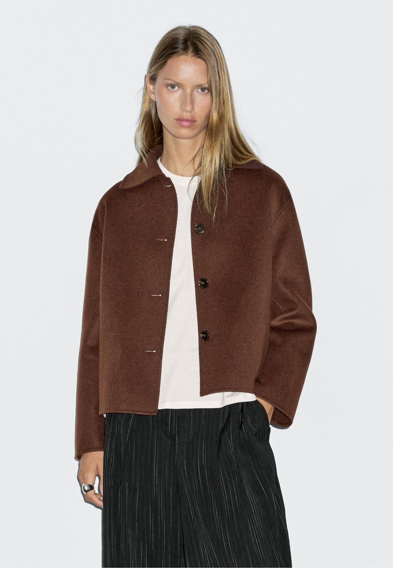 Giubbotto corto in lana marrone con colletto a punta, chiusura frontale con bottoni e una leggera lucentezza, abbinato a una camicia chiara e pantaloni con riga.