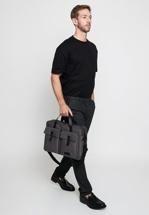 Mann, der mit einem grauen Aktkoffer geht, trägt ein schwarzes T-Shirt, graue Hose und schwarze Lederschuhe vor einem schlichten hellen Hintergrund.