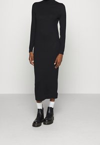 Schwarzes langärmeliges Kleid mit hohem Kragen, figurbetonter Silhouette und knielangem Saum. Kombiniert mit schwarzen Stiefeletten und weißen Knöchelsocken.