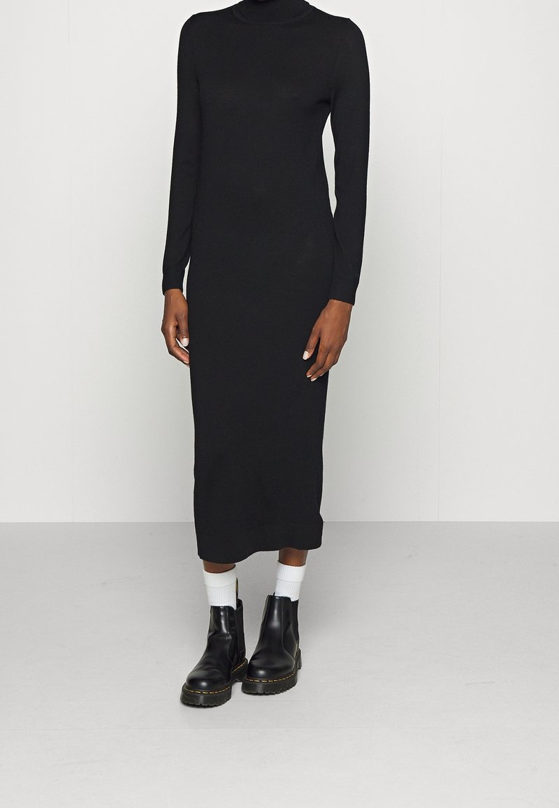 Schwarzes langärmeliges Kleid mit hohem Kragen, figurbetonter Silhouette und knielangem Saum. Kombiniert mit schwarzen Stiefeletten und weißen Knöchelsocken.