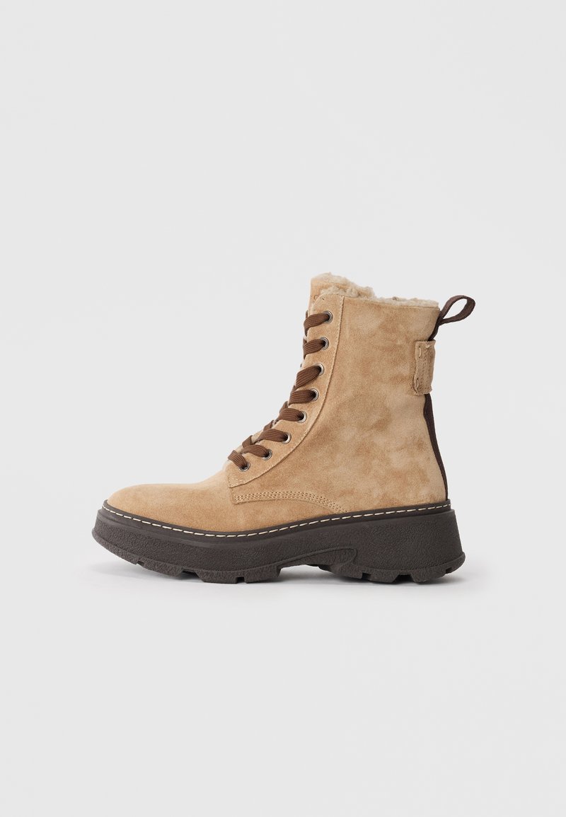 Tan suede lace-up ankle boot with thick black sole, padded collar, and pull tab on plain light background.