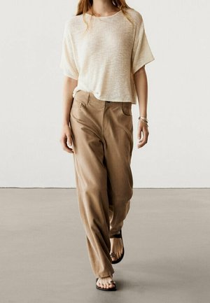 Femme marchant portant un top blanc ample à manches courtes, un pantalon large marron clair taille haute et des sandales noires à bouts ouverts.