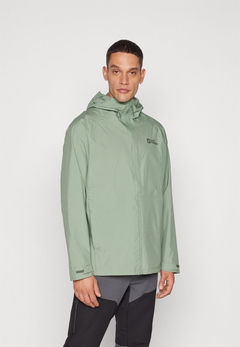Chaqueta impermeable verde claro con capucha, cremallera frontal y bolsillos laterales; presenta un logo en el pecho y una textura suave y mate.