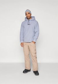 Gaiši zila kapuce ar auklu un logotipu, kopā ar bēšām biksēm ar kabatām. Melni zābaki noslēdz apģērbu. Vienkāršs, praktisks dizains.