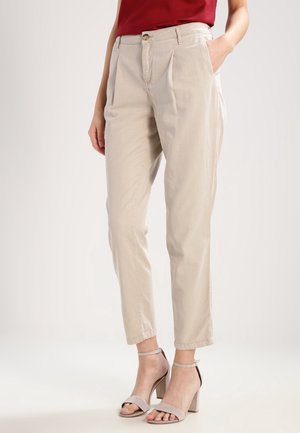 Femme portant un pantalon beige fuselé avec des plis, associé à des sandales taupe à talons blocs et bride à la cheville, et un haut rouge.