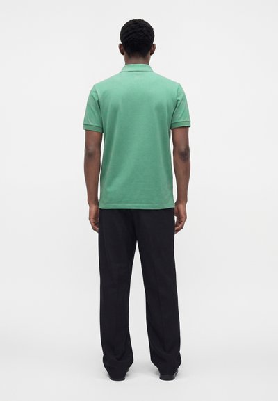 Homme debout de dos, portant un polo vert à manches courtes et un pantalon noir sur un fond blanc uni.