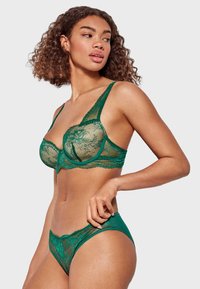 Sostén verde de encaje y tanga a juego, con patrones florales y paneles transparentes, con tirantes ajustables y bordes ondulados para más detalles.