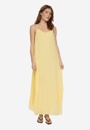 Kobieta ubrana w zwiewną, sięgającą kostek żółtą sukienkę na cienkich ramiączkach, stojąca i pozująca z rękami swobodnie opuszczonymi wzdłuż ciała.