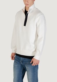 Sudadera blanca de manga larga con cremallera de un cuarto en el cuello, que presenta un cierre negro, puños acanalados y un ajuste holgado, combinada con jeans azules.
