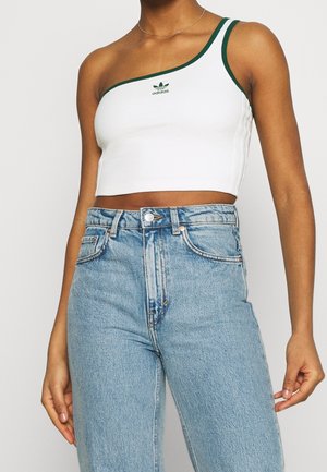 Kobieta ubrana w biały, jedno-ramienny crop top Adidas z zielonym wykończeniem i jasnoniebieskie, wysokiej talii dżinsy na jednolitym tle.