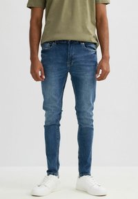 Jeans ajustados de mezclilla azul con desvanecimiento ligero, cinco bolsillos y cremallera. Combinados con una camiseta verde y zapatillas blancas.