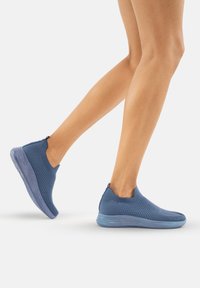 Zapatillas de lona azul sin cordones con parte superior texturizada, tacón sólido y suela translúcida, mostrando un diseño moderno y una construcción ligera.