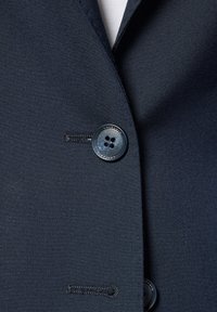 Szczegół marynarki z granatowego materiału, charakteryzującej się gładką fakturą, guzikiem z czterema otworami oraz kontrastującym czarnym przeszyciem po lewej stronie.