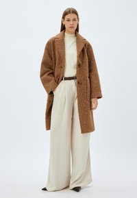Brun tekstureret frakke med store knapper, oversized pasform og brede revers, båret over en cremefarvet turtleneck og højtaljede beige bukser.