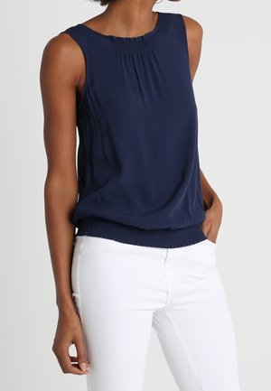Top sin mangas de color azul marino, hecho de una tela suave, con detalles fruncidos en el cuello y un dobladillo ligeramente elástico. Combinado con pantalones blancos.