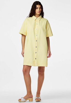 Naine seisab, kandes lühikeste varrukatega kollast ees nööbitavat kleiti ja valgeid kontsakingi tavalise valge tausta ees.