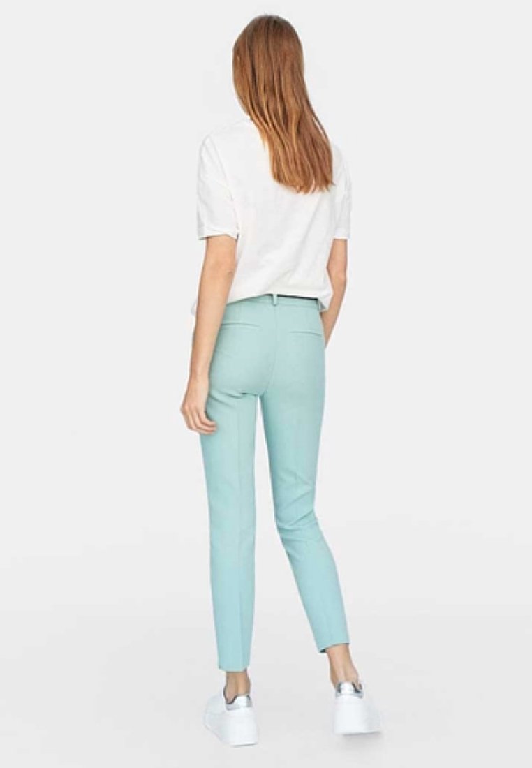 Femme aux cheveux longs châtain clair portant un t-shirt blanc à manches courtes, un pantalon slim bleu clair et des baskets blanches, debout de dos.