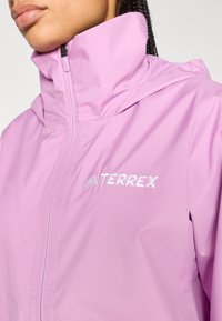 Veste imperméable violet clair avec col montant, fermeture éclair et logo Terrex sur la poitrine. Tissu lisse avec une légère brillance.