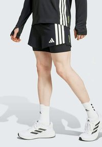 Pantalones cortos atléticos negros con rayas blancas, combinados con zapatos y calcetines blancos. El diseño incluye un logo de Adidas y una tela ligera.