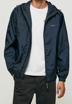 Mężczyzna ubrany w granatową, zapinaną na zamek kurtkę z kapturem, na białej koszulce i ciemnych spodniach, z rękami swobodnie opartymi przy bokach.