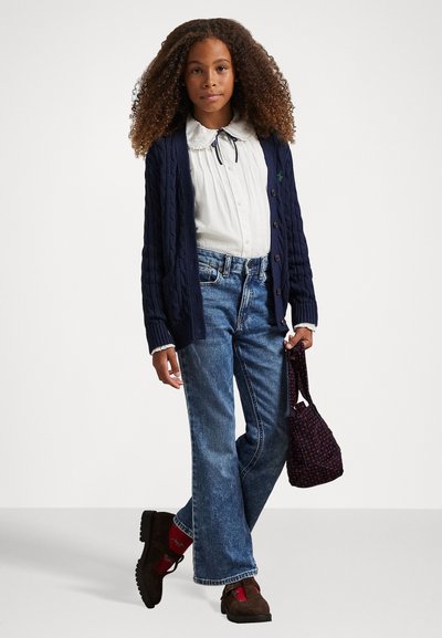 Young girl with curly hair wearing a navy cardigan, white blouse, blue jeans, brown shoes, red socks, holding a small patterned bag.