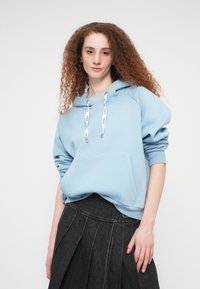 Γυναίκα με σγουρά κόκκινα μαλλιά, που φοράει ένα ανοιχτό γαλάζιο φούτερ με κουκούλα και σκουρόχρωμη πλισέ φούστα, στέκεται μπροστά σε απλό λευκό φόντο.