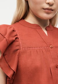 Femme portant un chemisier couleur rouille avec des manches courtes volantées, une bordure en dentelle sur le plastron et deux boutons visibles près de l'encolure.