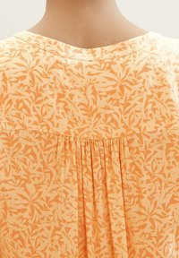 Blouse orange à motif de feuilles, avec un dos plissé, un col rond et un tissu doux et texturé.