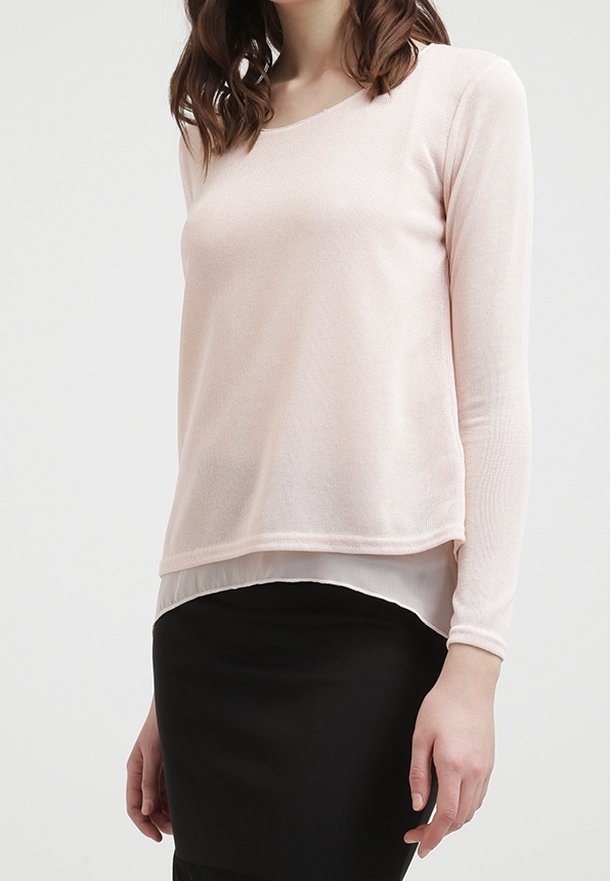 Femme portant un haut à manches longues rose clair à couches superposées, associé à une jupe noire ajustée, debout devant un fond uni.