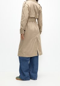 Kobieta w beżowym trenczu z paskiem, w szerokich niebieskich dżinsach i zielonych sneakersach, stojąca plecami do aparatu na białym tle.