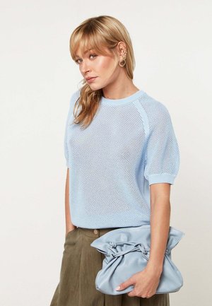 Femme aux cheveux blonds portant un haut en tricot bleu clair à manches courtes, une jupe olive, tenant une grande pochette plissée bleu clair.