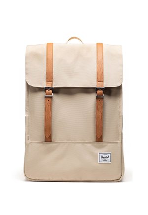 Beżowy prostokątny plecak z klapą, dwoma pionowymi brązowymi skórzanymi paskami z metalowymi klamrami oraz małą białą naszywką z logo u dołu.