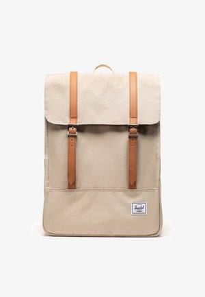 Beige rectangular backpack with flap closure, two vertical brown leather straps with metal buckles, and a small white brand patch at the bottom.