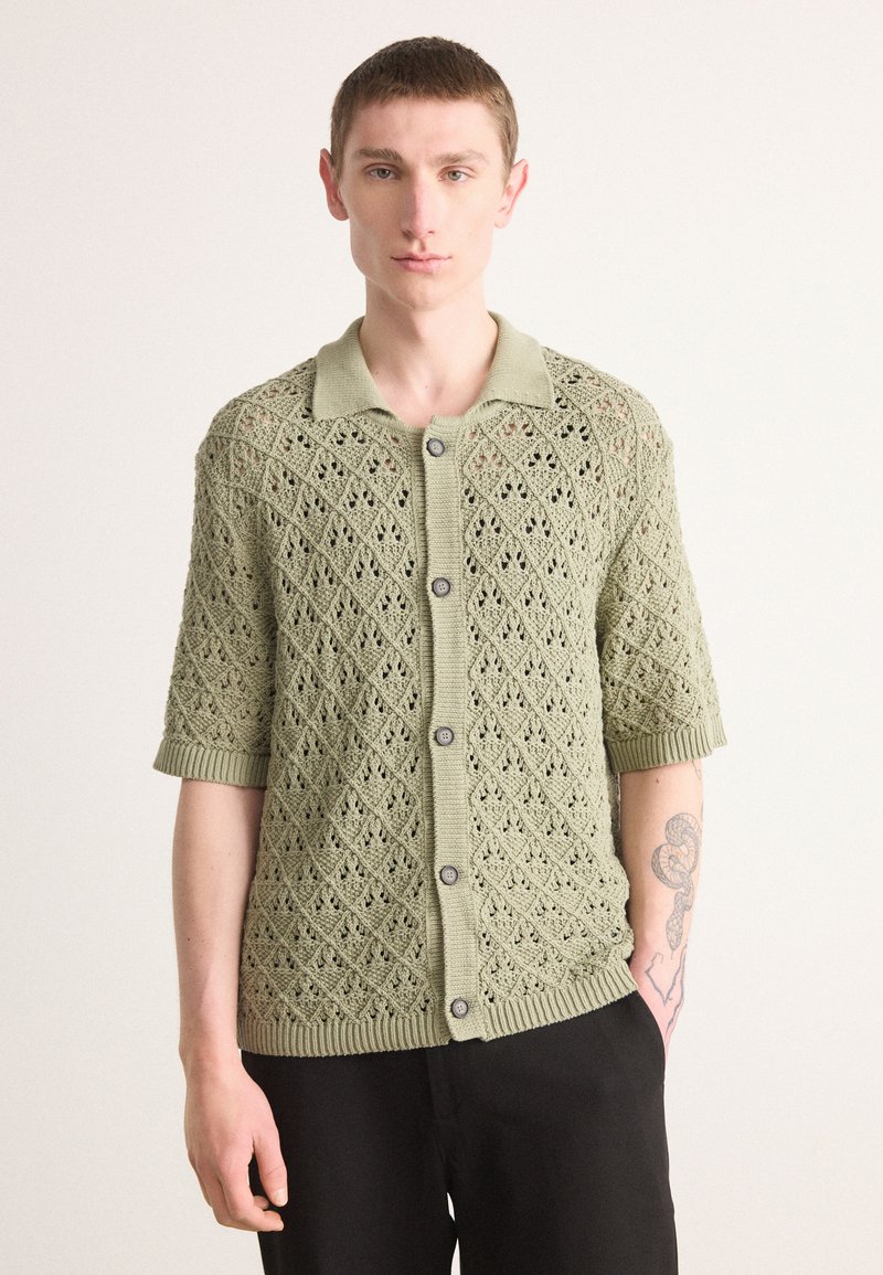 Helle grüne Strickjacke mit Rautenmuster, kurzen Ärmeln und geripptem Kragen. Fünf dunkle Knöpfe vorne.