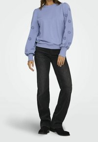 Kvinna i en lavendellila långärmad topp med blommönster på ärmarna, svarta raka jeans och svarta stövlar, stående mot en vit bakgrund.