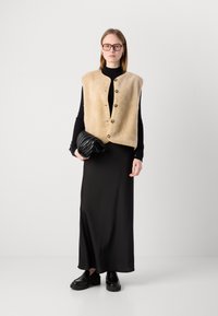Beige pälsväst med knappstängning, svart turtleneck, svart maxikjol och platåskor. Modellen håller en viks svart clutch.