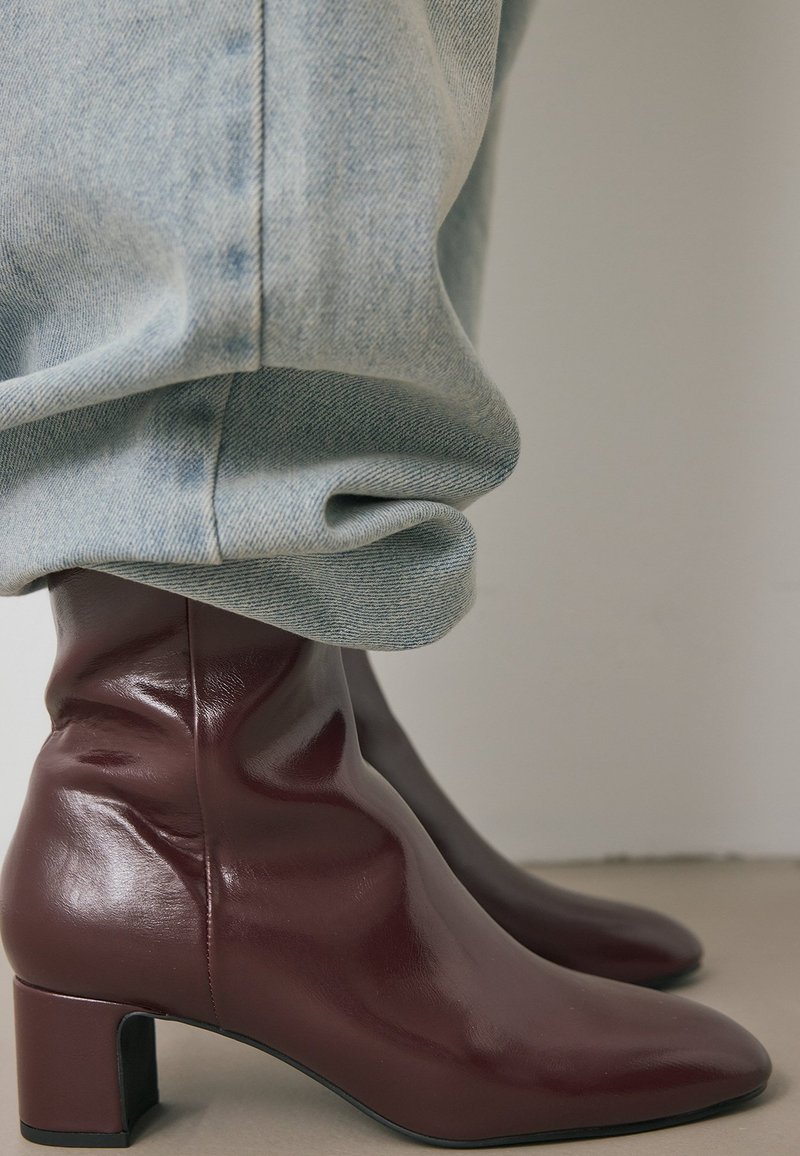 Botas de cuero burdeos hasta el tobillo con un tacón alto y cuadrado; textura suave y diseño elegante, combinadas con pantalones de mezclilla azul claro.