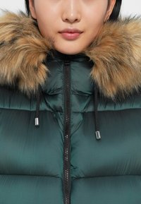 Chaqueta inflable verde con cuello de piel, que presenta un cierre de cremallera y cordones. El material parece suave, con secciones acolchadas y herrajes plateados.
