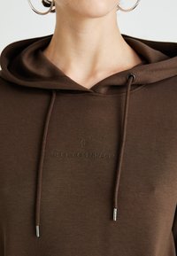 Detailní záběr na osobu v hnědé mikině s kapucí a šňůrkami, na které je logo "MSCH Copenhagen" na hrudi, a má také stříbrné kruhové náušnice.