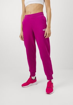 Woman wearing bright pink Puma sweatpants and matching sneakers, standing against a plain white background, cropped at the waist.