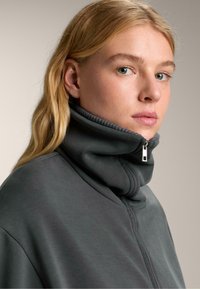 Šedá mikina na zips s vysokým golierom. Mäkký materiál, štruktúrovaný dizajn a strieborný zips pri krku. Hladká, textúrovaná tkanina.