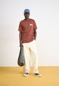 T-shirt marron à manches courtes avec le logo 'Dickies', pantalon large crème, baskets noires et casquette en denim bleu. Tenant un sac à motifs.