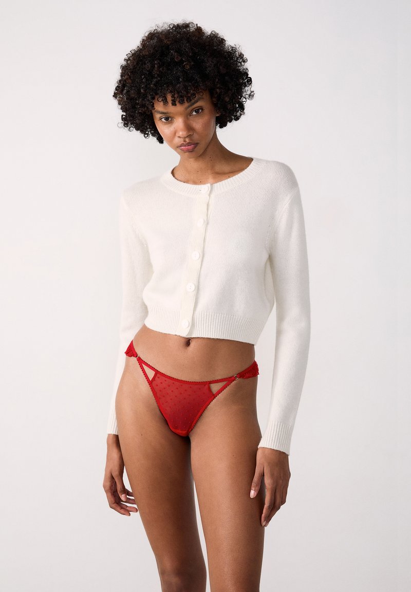 Cardigan bianco lavorato a maglia con maniche lunghe e bottoni, abbinato a un perizoma rosso traslucido con pois e rifiniture in pizzo e dettagli con tagli.