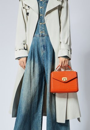 Borsa a tracolla arancione con stampa a coccodrillo e chiusura in oro, tenuta da una persona che indossa un trench grigio chiaro con cintura e una tuta in denim.