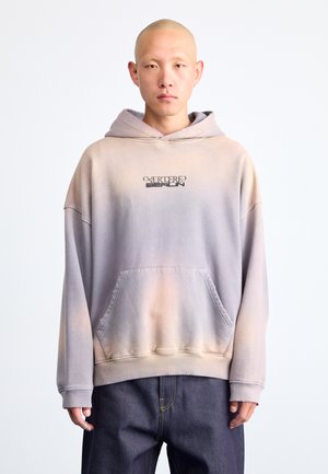 Oversized hoodie in gradient grey to peach hues, made of soft fabric, with a front pocket and black logo text. Features ribbed cuffs.
