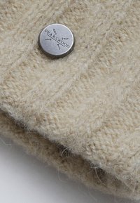 Beige pletený šátek s jemnou strukturou. Obsahuje stříbrný kovový knoflík s vyraženým nápisem "PEARLWOOD." Jednoduchý design s útulným vzhledem.