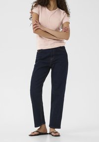 Woman standing with arms crossed, wearing a light pink t-shirt, dark blue jeans, and black flip-flops on a plain background.